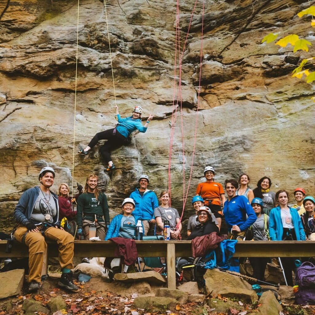 Group of climbers in helmets and harnesses pose together at the base of a rock wall, one person mid-climb on the wall above.