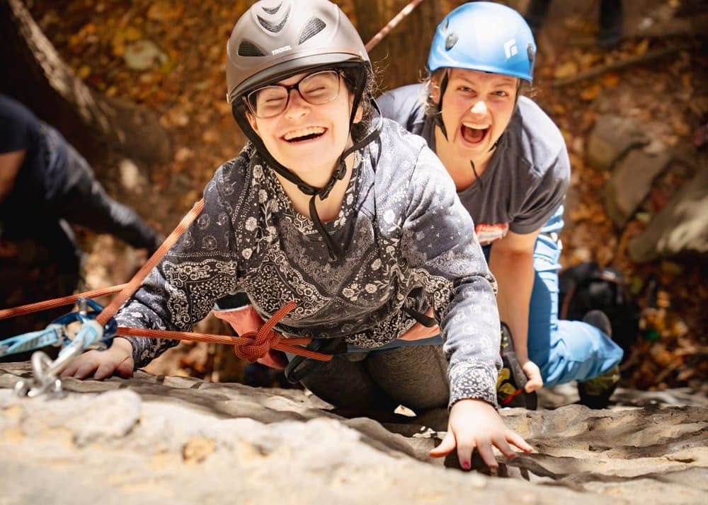 Two young climbers wearing helmets rock climbing outdoors, one smiling at the camera as the other cheers behind.