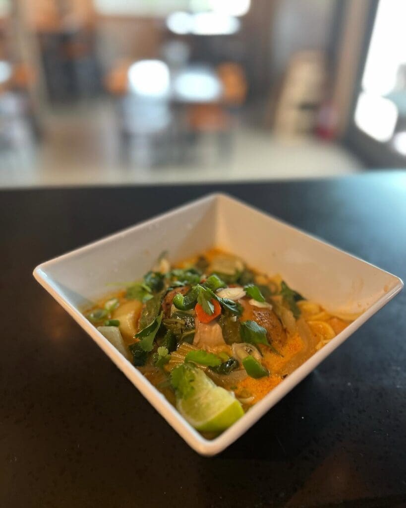 Square white bowl of spicy soup with herbs, vegetables, and a lime wedge on a dark table; blurred restaurant background behind.