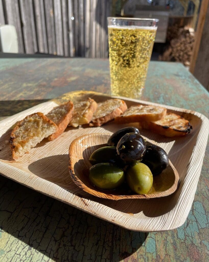 Wooden tray with sliced baguette and a bowl of green and black olives on a rustic table, a cold glass of beer in the background.