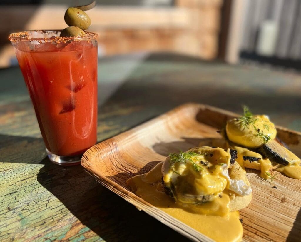 Tall glass of red tomato cocktail with olive garnish and a salted rim, beside a wooden board of eggs Benedict with hollandaise.