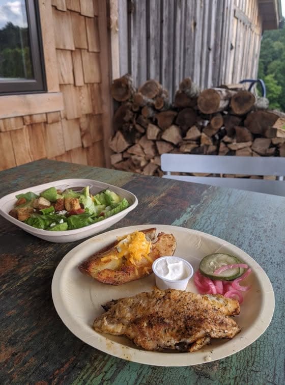 Outdoor rustic setting with a plate of grilled chicken, salad bowl, and toppings on a weathered table; stacked firewood and wooden siding in the background.
