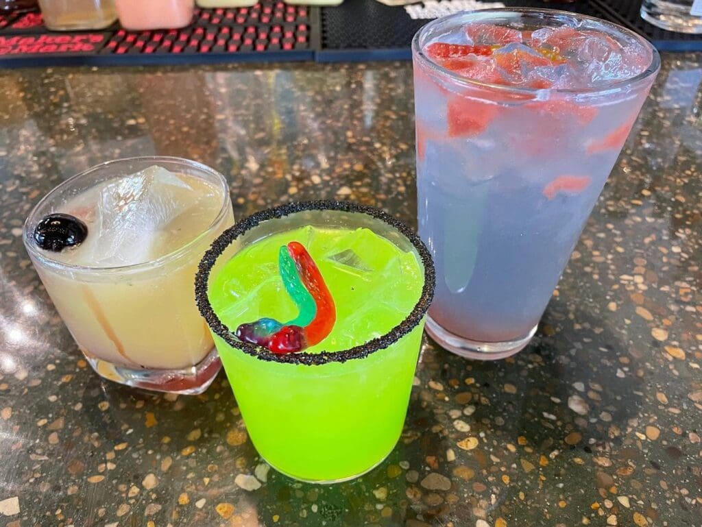 Three colorful cocktails on a speckled bar: neon green drink with a black sugar rim and gummy snake, pale yellow drink with a cherry, and a tall blue beverage with ice and fruit.
