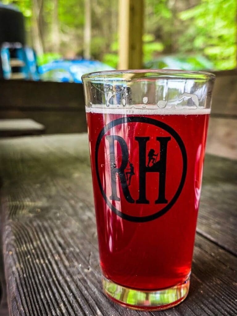 Pint glass of red beer on a wooden outdoor table with a black circular logo and small climber silhouettes inside the logo.