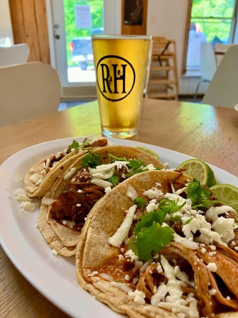 Plate of soft tacos with shredded meat, cheese, cilantro and lime wedges on a wooden table; a tall beer glass with an RH logo in the background.
