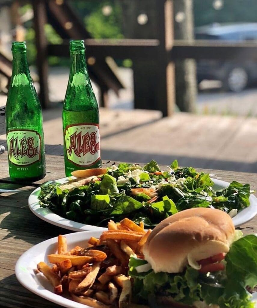 Two green Ale8 bottles sit on a sunny wooden table beside plates of a burger, fries, and a large leafy salad outdoors.