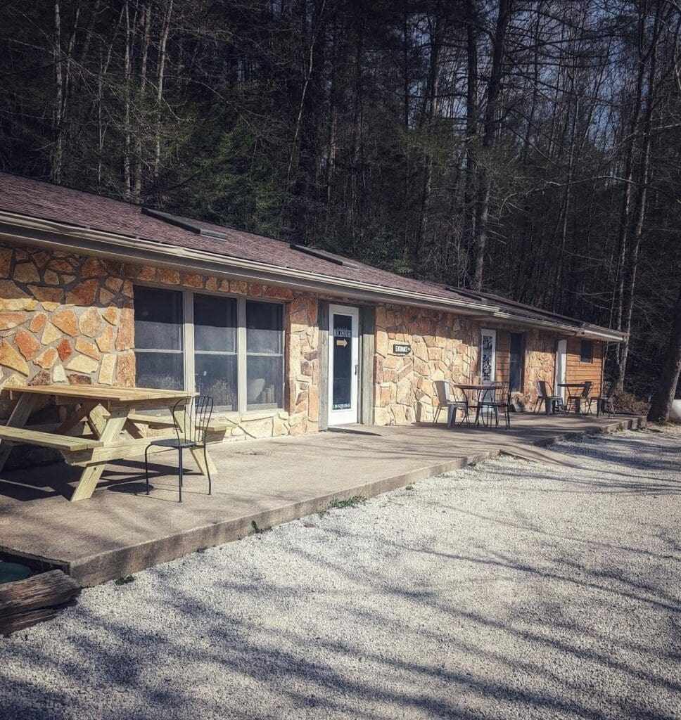 Stone building with a gravel lot and outdoor seating (tables and chairs) along a woodsy backdrop of trees.