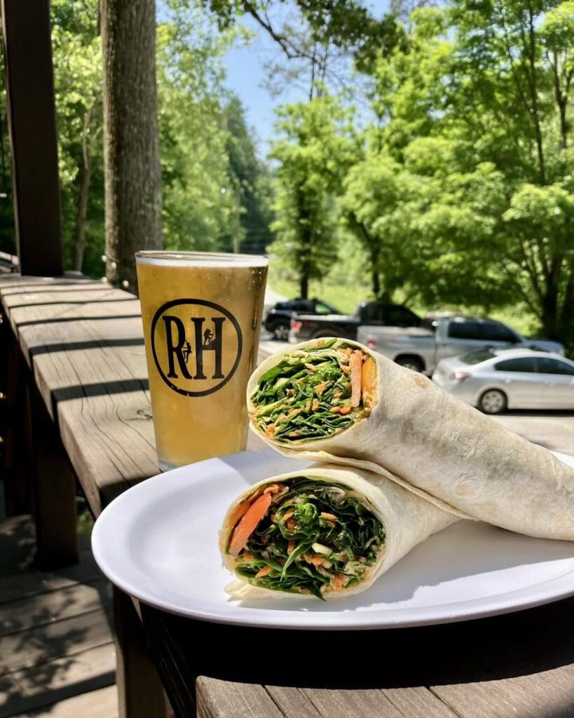 Two veggie wraps on a white plate with a cold glass of beer outdoors on a wooden railing, surrounded by green trees.