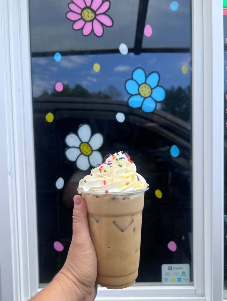 Hand holding a iced coffee with whipped cream and colorful sprinkles in front of a window with flower decals.