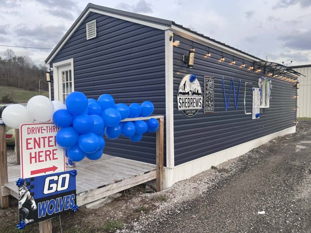 Blue coffee trailer with white trim, festive blue balloons, string lights, and signs reading SHEBREWS and WOLVES outside.