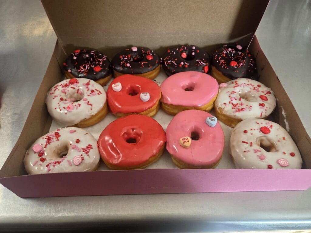 Box of assorted donuts with white, pink, and red icing and pink sprinkles in a cardboard bakery box