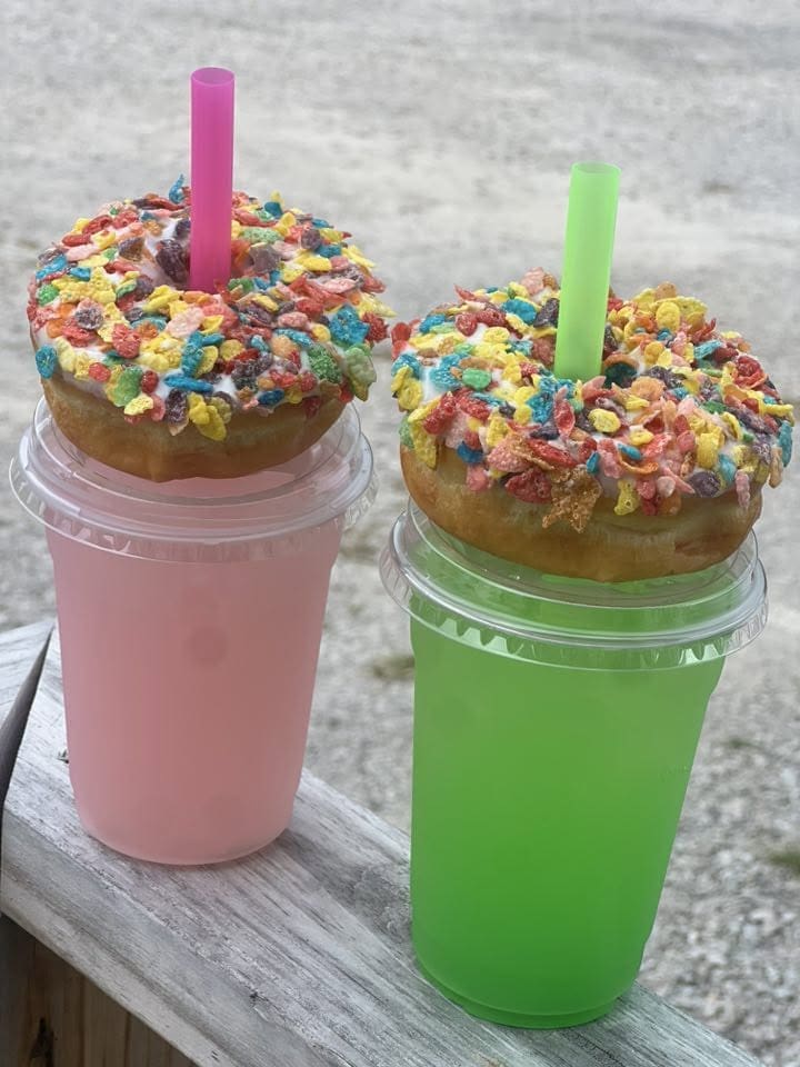Two colorful smoothie cups topped with sprinkled donuts and large pink and green straws, resting on a wooden railing outdoors.