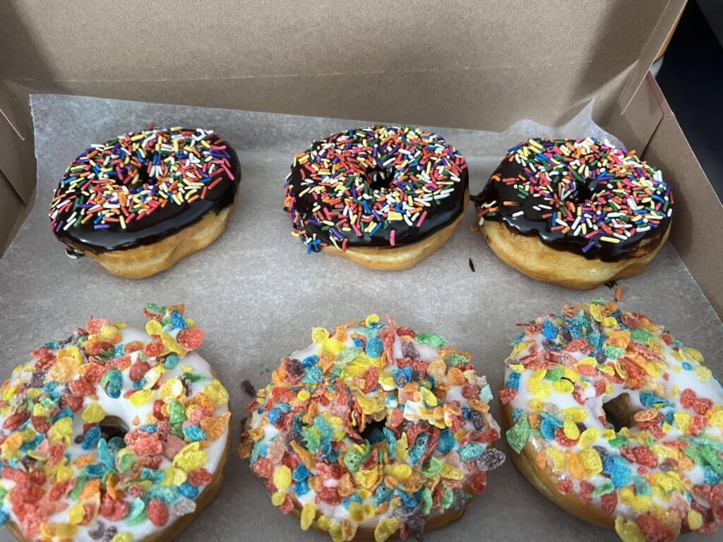 Three donuts with chocolate glaze and colorful rainbow sprinkles sit in an open cardboard box.